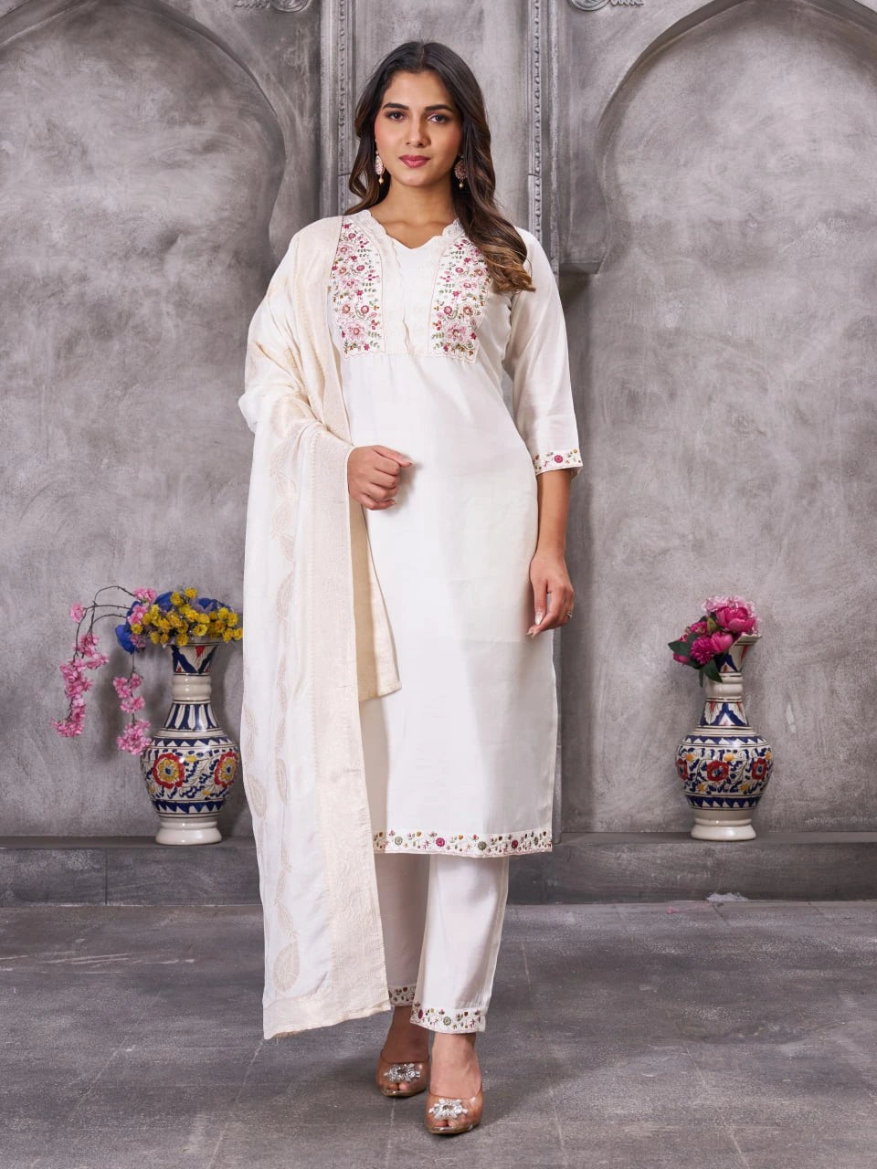 Woman in a white traditional outfit with floral embroidery standing against a gray wall with decorative vases.